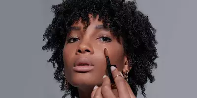 Mujer con tono de piel oscura que muestra cómo aplicar el corrector All Over debajo de los ojos.